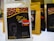 A package of Aman Kuba Luwak coffee is prominently displayed in the foreground, featuring a graphic of coffee beans and gold text on a black background. Other similar coffee packages are arranged in the background, slightly blurred.