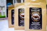 Three brown paper bags of specialty coffee are arranged on a tiled surface. Each bag features a label with a circular design, including the text 'Specialty Coffee' and '100% Pure Arabica'. There is detailed artwork involving a landscape and some text in both English and possibly another language. In the background, a water dispenser is visible.