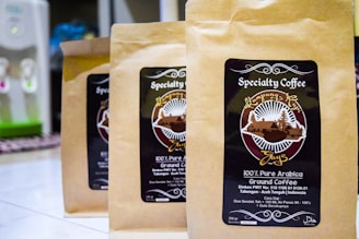 Three brown paper bags of specialty coffee are arranged on a tiled surface. Each bag features a label with a circular design, including the text 'Specialty Coffee' and '100% Pure Arabica'. There is detailed artwork involving a landscape and some text in both English and possibly another language. In the background, a water dispenser is visible.