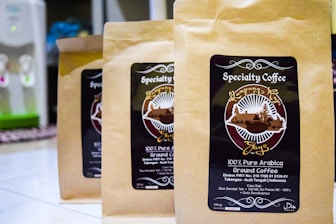 Three brown paper bags of specialty coffee are arranged on a tiled surface. Each bag features a label with a circular design, including the text 'Specialty Coffee' and '100% Pure Arabica'. There is detailed artwork involving a landscape and some text in both English and possibly another language. In the background, a water dispenser is visible.
