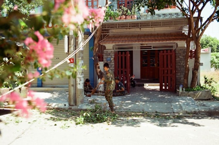 A residential house with a red gate and lush foliage in the foreground. A person is sweeping or cleaning leaves off the pavement. Another person can be seen sitting near the gate, possibly resting. The scene includes vibrant pink flowers partially obscuring the view, and the area is shaded by trees.