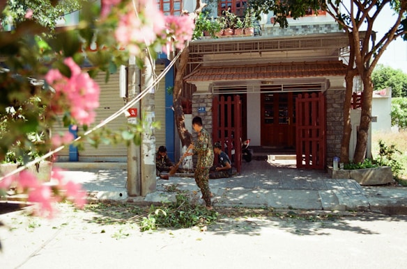 A residential house with a red gate and lush foliage in the foreground. A person is sweeping or cleaning leaves off the pavement. Another person can be seen sitting near the gate, possibly resting. The scene includes vibrant pink flowers partially obscuring the view, and the area is shaded by trees.