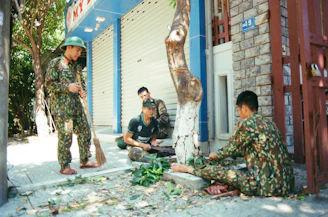 Four people wearing camouflage uniforms are working together outdoors. They appear to be cleaning up a sidewalk area, with one person sweeping while others are sitting near a tree, possibly picking up leaves or debris. The closed shutters of a building are in the background, and a placard with the number '19' is visible on the wall.