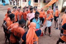 A group of people wearing matching orange t-shirts and bandanas, gathered in a semi-circle. Some of them are playing musical instruments such as drums, while others hold flags. They appear to be engaged in a celebratory or cultural activity in an outdoor setting with buildings visible in the background.