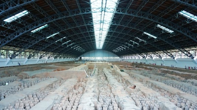 A large indoor archaeological excavation site housing a vast number of ancient clay statues, arranged in neat rows. The high ceiling, supported by a robust metal framework, allows ample natural light to illuminate the statues. A long walkway on the right side is occupied by visitors observing the scene.