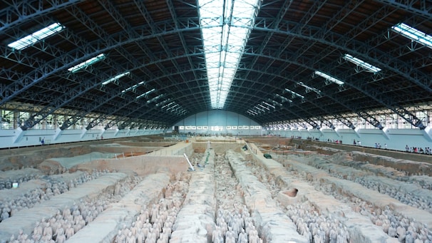 A large indoor archaeological excavation site housing a vast number of ancient clay statues, arranged in neat rows. The high ceiling, supported by a robust metal framework, allows ample natural light to illuminate the statues. A long walkway on the right side is occupied by visitors observing the scene.