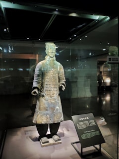 A Terracotta Warrior statue stands inside a glass display case. The figure is dressed in detailed armor, with a stern expression and hands positioned as if holding something. The lighting casts dramatic shadows, highlighting the intricate craftsmanship. Adjacent to the statue is an informational plaque with text in both Chinese and English.