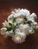 A bouquet of white flowers, including roses, tulips, and peonies, resting on a wooden surface. The flowers are fresh and lush, with green leaves interspersed among the blossoms, creating a natural and elegant arrangement.