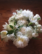 A bouquet of white flowers, including roses, tulips, and peonies, resting on a wooden surface. The flowers are fresh and lush, with green leaves interspersed among the blossoms, creating a natural and elegant arrangement.