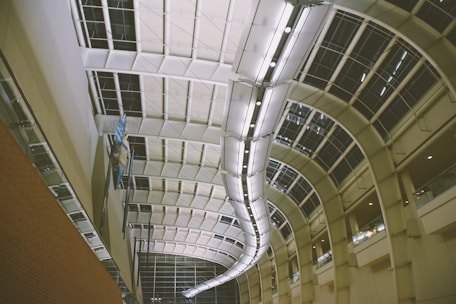 An architectural scene featuring a large indoor atrium with a high, curved ceiling composed of metal frames and glass panels. Fluorescent lights run along the spine of the ceiling, creating a bright, airy space. The view shows a contrasting brick wall on the left side and open balconies overlooking the atrium.