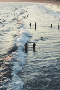 People are enjoying the ocean along a beach, with individuals scattered throughout the water as waves roll in. The scene offers a sense of leisure and relaxation, with the sun casting a warm glow over the scene.