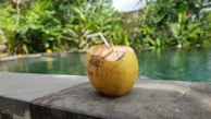 A fresh coconut with a straw is placed on a concrete ledge by a swimming pool, surrounded by lush green foliage.