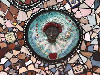 A colorful mosaic featuring a central circular piece with a three-dimensional face surrounded by vibrant tiles. The face has red-like adornments around the forehead and is set against a teal background with small geometric and floral patterns. The text 'REINA DE LA MANZANAS' is visible around the face. Surrounding the central circle are various irregularly shaped tiles in colors like blue, pink, brown, and white, creating a patchwork effect.