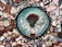 A colorful mosaic featuring a central circular piece with a three-dimensional face surrounded by vibrant tiles. The face has red-like adornments around the forehead and is set against a teal background with small geometric and floral patterns. The text 'REINA DE LA MANZANAS' is visible around the face. Surrounding the central circle are various irregularly shaped tiles in colors like blue, pink, brown, and white, creating a patchwork effect.
