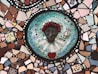 A colorful mosaic featuring a central circular piece with a three-dimensional face surrounded by vibrant tiles. The face has red-like adornments around the forehead and is set against a teal background with small geometric and floral patterns. The text 'REINA DE LA MANZANAS' is visible around the face. Surrounding the central circle are various irregularly shaped tiles in colors like blue, pink, brown, and white, creating a patchwork effect.