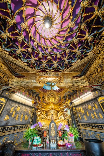 An ornate temple interior featuring an intricate ceiling design with a radiant light at the center. The foreground displays a golden statue of a deity surrounded by numerous smaller arms, symbolizing multiplicity or compassion. Below the statue, offerings, including flowers and incense, are placed on an altar.