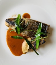 A gourmet dish featuring a piece of grilled fish with crispy skin, garnished with green herbs and served alongside vegetables. The plate includes a rich brown sauce and a smooth, creamy puree, artistically arranged on a white plate.