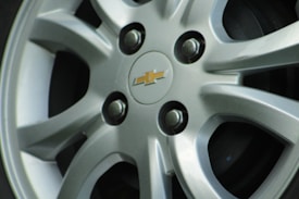A close-up of a car wheel with a five-spoke alloy rim featuring a Chevrolet logo in the center. The rims are silver with a smooth, polished finish, and the bolts are visible and securely fastened around the center cap.
