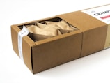 A rectangular cardboard box partially opened to reveal its contents, which include multiple paper-wrapped items. The box features a white label on top with text and a small design element.