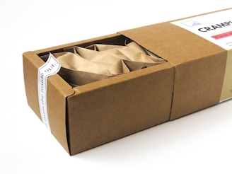 A rectangular cardboard box partially opened to reveal its contents, which include multiple paper-wrapped items. The box features a white label on top with text and a small design element.
