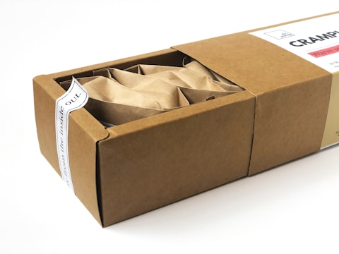 A rectangular cardboard box partially opened to reveal its contents, which include multiple paper-wrapped items. The box features a white label on top with text and a small design element.