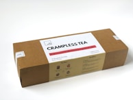 A rectangular cardboard box labeled 'CRAMPLESS TEA' by 'dr. yang', which is described as a warm soothing blend for period pain. The box contains 10 wrapped tea bags and specifies that the tea is caffeine-free and a herbal supplement. The design includes instructions on usage and benefits labeled 'The How' and 'The When' on the package.