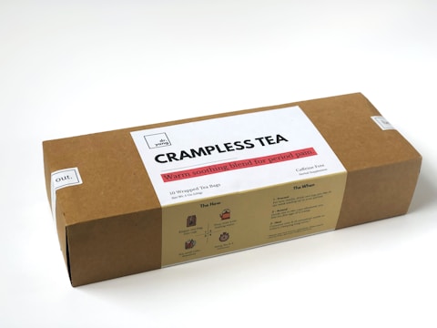 A rectangular cardboard box labeled 'CRAMPLESS TEA' by 'dr. yang', which is described as a warm soothing blend for period pain. The box contains 10 wrapped tea bags and specifies that the tea is caffeine-free and a herbal supplement. The design includes instructions on usage and benefits labeled 'The How' and 'The When' on the package.