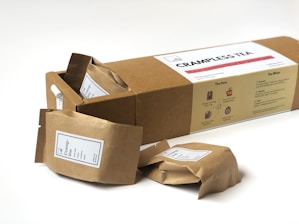 A box labeled 'CRAMPLESS TEA' accompanied by several brown paper packets with white labels. The background is plain and the focus is on the tea packaging. The box includes instructions for brewing tea.