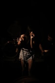 A female singer stands on stage, gripping a microphone with both hands. She is wearing a dark top, denim shorts, and fishnet stockings. The scene is dimly lit, casting shadows on her and the musicians behind her, creating a moody atmosphere.