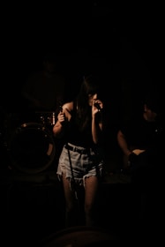 A female singer stands on stage, gripping a microphone with both hands. She is wearing a dark top, denim shorts, and fishnet stockings. The scene is dimly lit, casting shadows on her and the musicians behind her, creating a moody atmosphere.