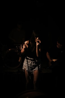 A female singer stands on stage, gripping a microphone with both hands. She is wearing a dark top, denim shorts, and fishnet stockings. The scene is dimly lit, casting shadows on her and the musicians behind her, creating a moody atmosphere.