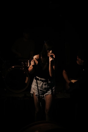 A female singer stands on stage, gripping a microphone with both hands. She is wearing a dark top, denim shorts, and fishnet stockings. The scene is dimly lit, casting shadows on her and the musicians behind her, creating a moody atmosphere.