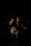 A female singer stands on stage, gripping a microphone with both hands. She is wearing a dark top, denim shorts, and fishnet stockings. The scene is dimly lit, casting shadows on her and the musicians behind her, creating a moody atmosphere.