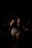 A female singer stands on stage, gripping a microphone with both hands. She is wearing a dark top, denim shorts, and fishnet stockings. The scene is dimly lit, casting shadows on her and the musicians behind her, creating a moody atmosphere.