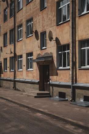 A multi-story residential building with a worn, textured facade. Several satellite dishes are mounted on the exterior, and metal pipes run vertically along the building. The windows are uniform, with white frames, and the entrance has a small set of steps leading up to a wooden door.