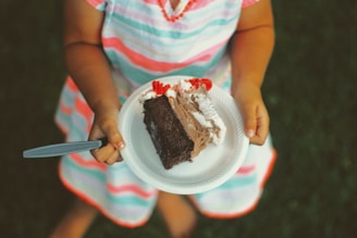 A child wearing a colorful striped dress holds a plate with a slice of chocolate cake topped with whipped cream and cherries. The child grips a fork ready to enjoy the dessert while standing on grass.