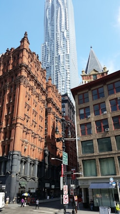 A bustling urban scene featuring a mix of architectural styles. A modern skyscraper towers in the background, showcasing a sleek and futuristic design. In the foreground, there are older brick buildings with ornate detailing, reflecting classic architecture. Pedestrians and street signs are visible on the busy city streets below.