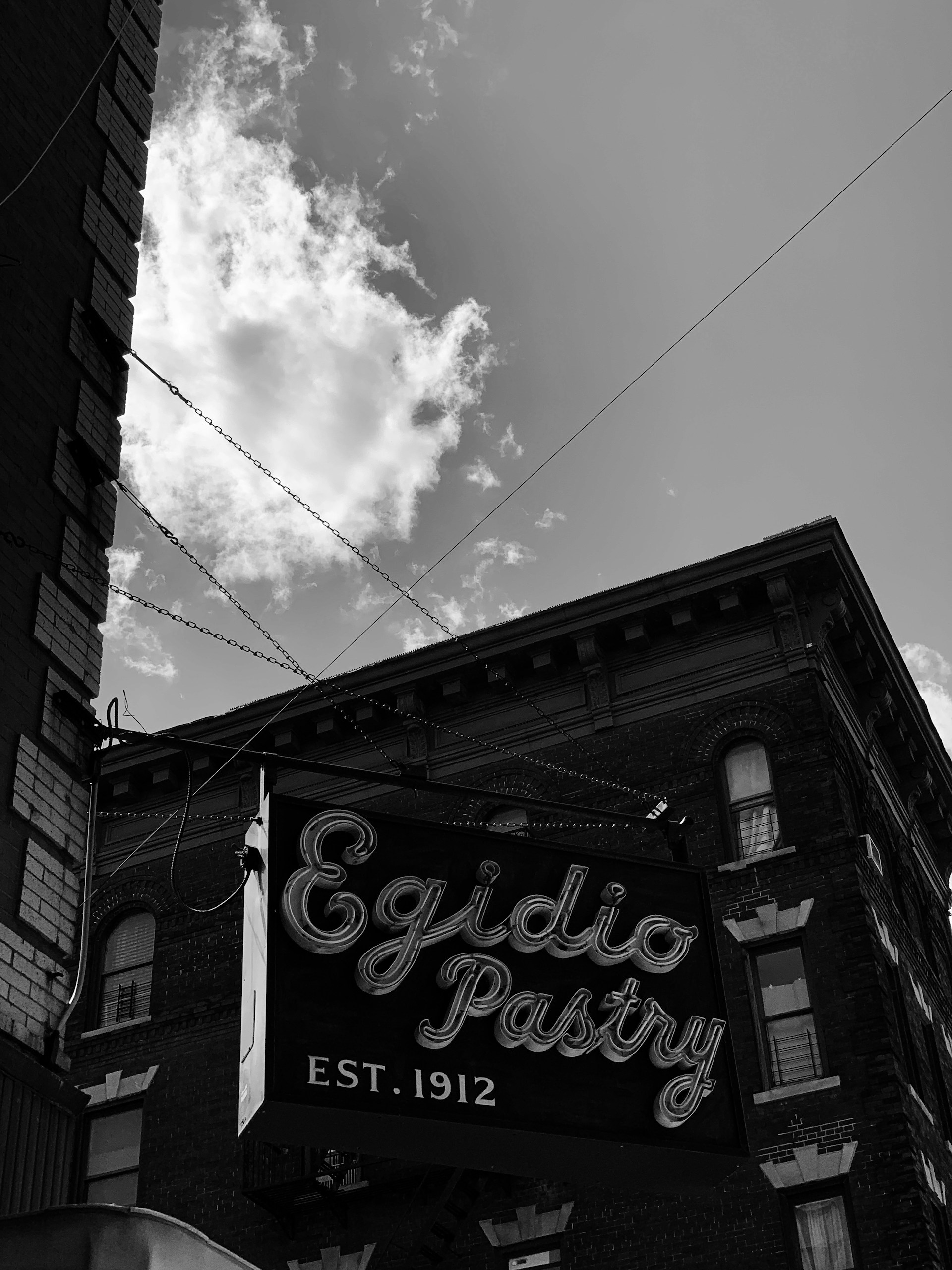 A vintage-style sign for 'Egidio Pastry' with 'Est. 1912' written on it hangs in front of a brick building. The sign is against a backdrop of a partly cloudy sky.