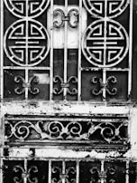 An ornate metal gate featuring circular and swirling patterns, with a weathered and rustic appearance. The intricate designs suggest traditional or cultural influences. The metal appears to be black and white due to an artistic, high-contrast black-and-white treatment.