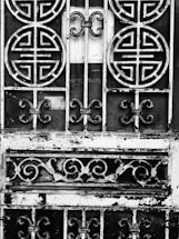 An ornate metal gate featuring circular and swirling patterns, with a weathered and rustic appearance. The intricate designs suggest traditional or cultural influences. The metal appears to be black and white due to an artistic, high-contrast black-and-white treatment.
