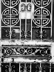 An ornate metal gate featuring circular and swirling patterns, with a weathered and rustic appearance. The intricate designs suggest traditional or cultural influences. The metal appears to be black and white due to an artistic, high-contrast black-and-white treatment.