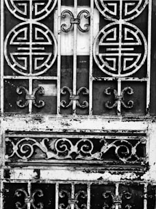 An ornate metal gate featuring circular and swirling patterns, with a weathered and rustic appearance. The intricate designs suggest traditional or cultural influences. The metal appears to be black and white due to an artistic, high-contrast black-and-white treatment.