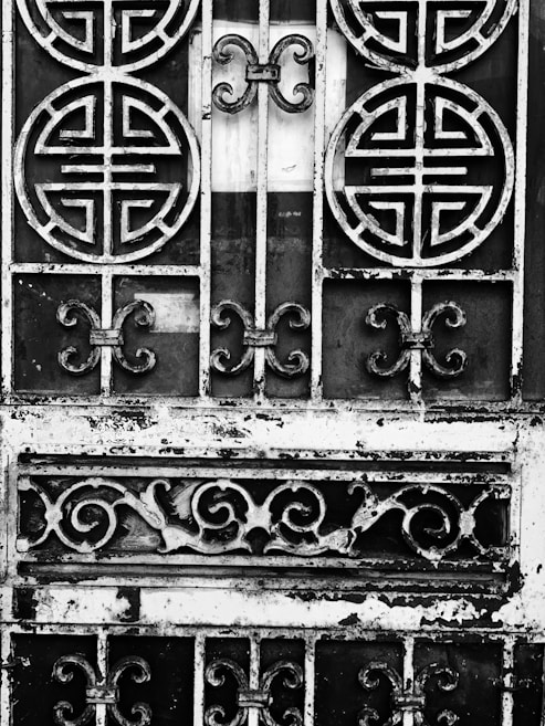 An ornate metal gate featuring circular and swirling patterns, with a weathered and rustic appearance. The intricate designs suggest traditional or cultural influences. The metal appears to be black and white due to an artistic, high-contrast black-and-white treatment.