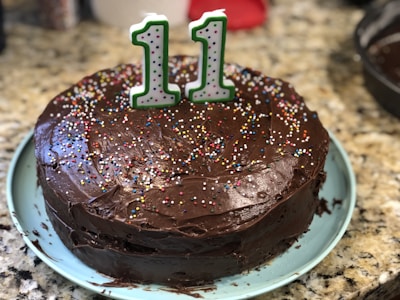 A chocolate cake with brown frosting is topped with colorful sprinkles and a numeral candle displaying the number 11. The cake is placed on a light blue plate, which is set on a granite countertop.