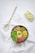 A healthy bowl filled with various ingredients including chickpeas, green peas, corn, avocado, lettuce, red hummus, and flaxseeds. The bowl is set on a white fabric with a wooden spoon on the side. Nearby, there are lemon wedges on a small white dish.