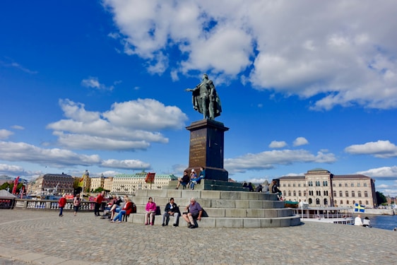 A large bronze statue of a historical figure stands prominently on a stone pedestal in a public square. People are sitting and walking around the monument. The background features classic European architecture with several buildings under a bright blue sky scattered with fluffy white clouds.