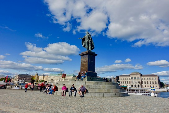 A large bronze statue of a historical figure stands prominently on a stone pedestal in a public square. People are sitting and walking around the monument. The background features classic European architecture with several buildings under a bright blue sky scattered with fluffy white clouds.