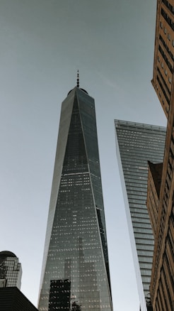 A towering skyscraper with a sleek, modern design rises towards a clear sky, flanked by other tall buildings. The glass facade reflects the surroundings, contributing to the grandeur and urban feel.