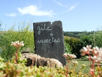 A slate sign with the words 'gite &agrave; insectes' stands in a lush garden setting, surrounded by various green plants and flowers. In the background, rolling hills and a clear blue sky create a serene and natural environment.