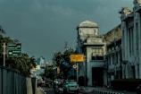 A street scene featuring an urban environment with historical architecture on the right side, including a building with a clock tower. Several vehicles and motorbikes are on the road, and traffic signs in Indonesian are visible, including one cautioning about a busway.